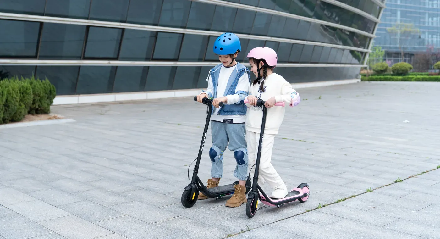 Deux enfants casqués et protégés aux genoux, debout sur deux trottinettes Evercross E6, noire et rose, sur une esplanade pavée