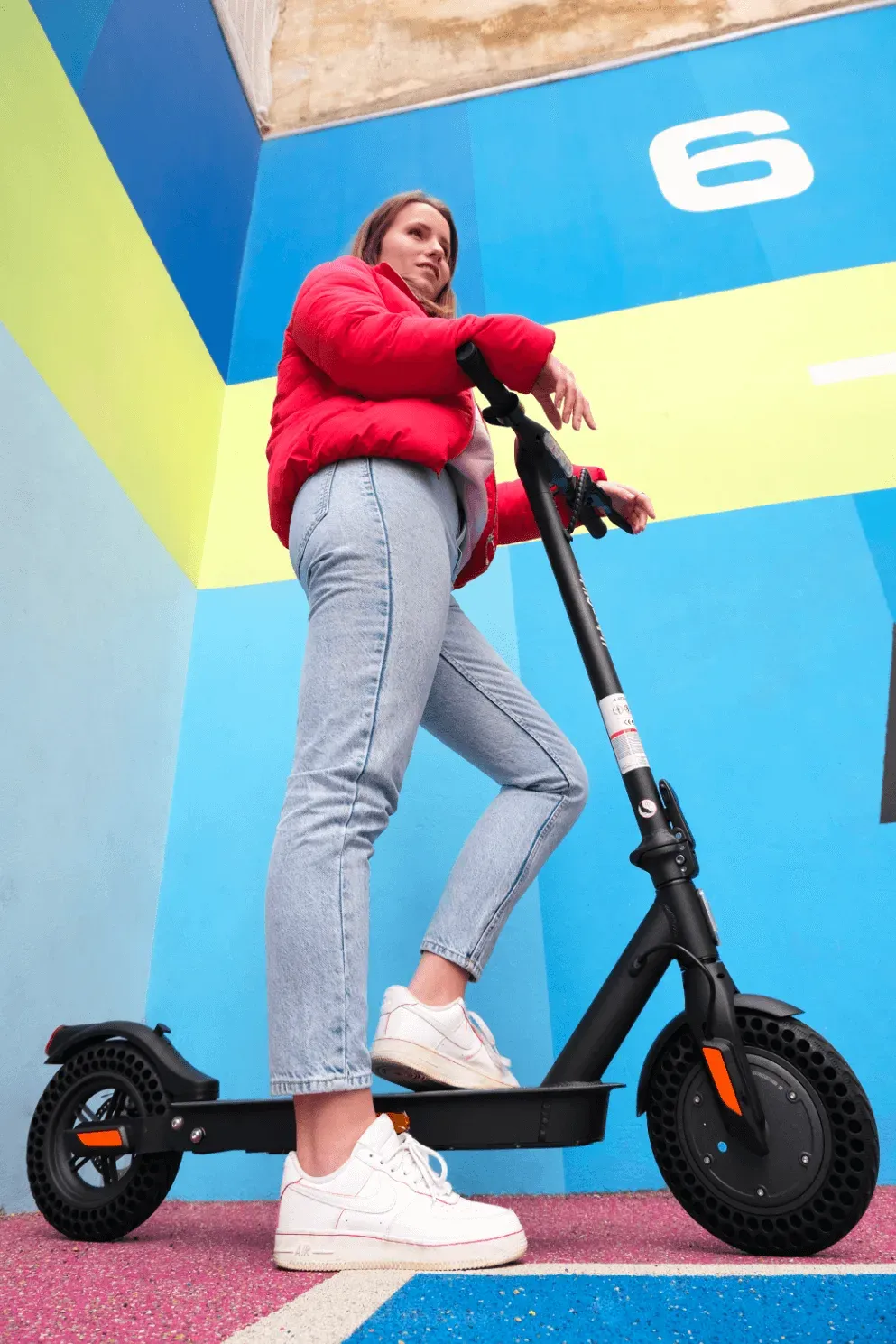 Une jeune femme debout sur l'Urbanglide Ride 100, un pied sur le plateau, devant un mur peint en bleu